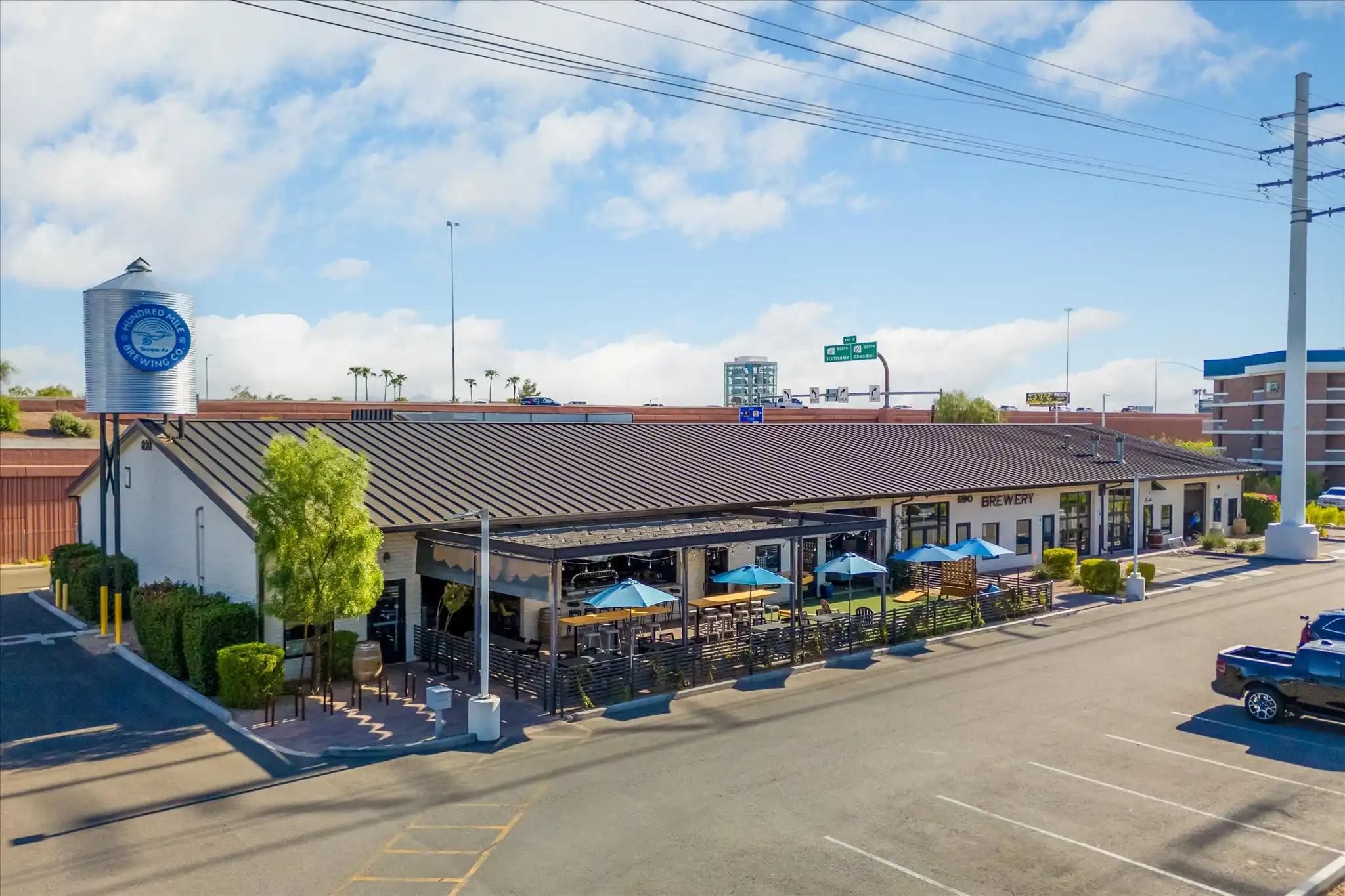 Evening on the Hundred Mile patio, with string lights, a wood pergola, blue umbrellas, and the brewery sign.